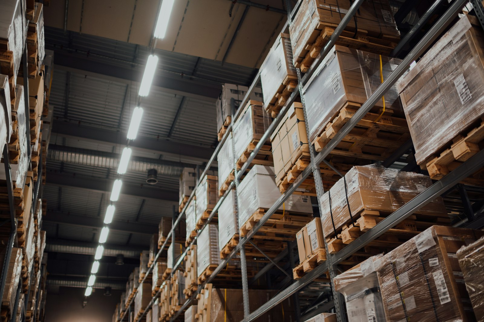 Pallets of boxed goods on tall warehouse racking under strip lights